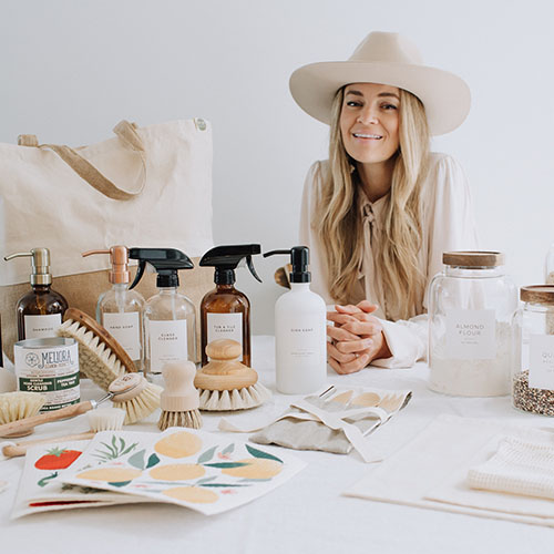 Kimberly Pymm, a white woman wearing a cream hat and shirt, sits at a table filled with bottles, jars, and other waste-free products. She is leaning on the table and smiling.