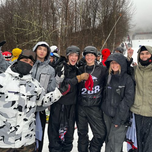 6 smiling students from the ski and snowboard team standing at the base of a snowy ski run