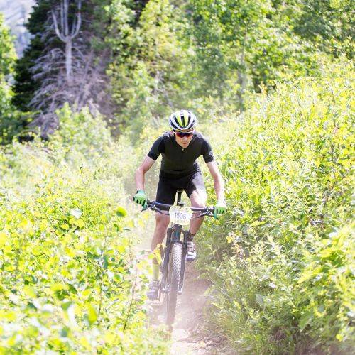 a man wearing black mountain biking down a trail with green plants on each side