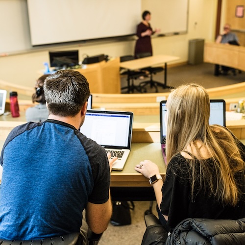 Students taking notes on laptops in a tiered lecture hall while an instructor teaches at the front.