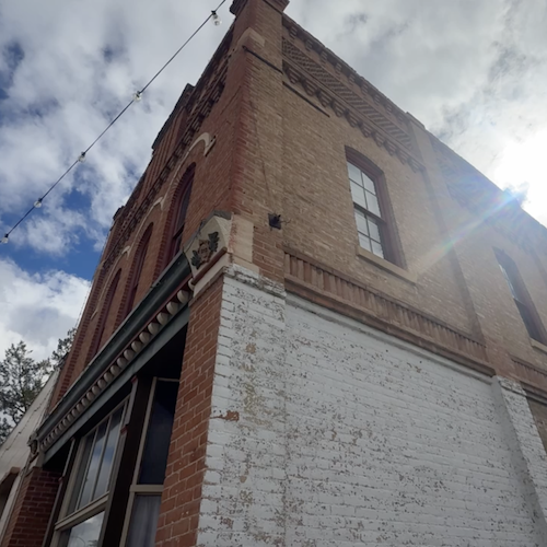 Low-angle view of a brick corner building with a white-painted lower facade and sun glare in a cloudy sky.