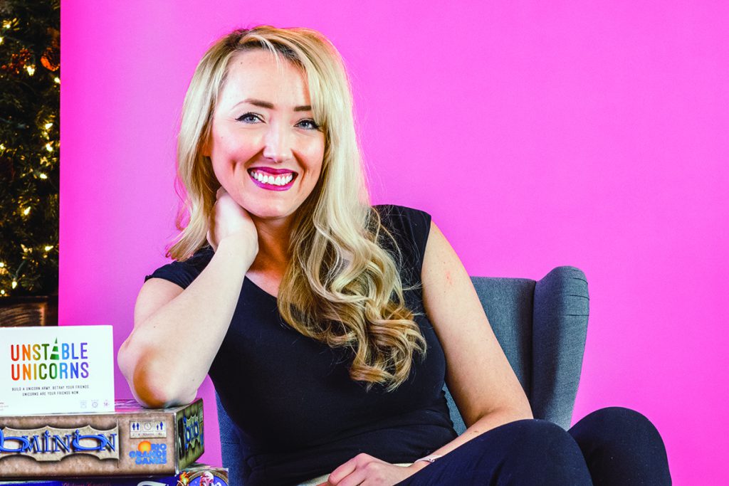 Erin Fair leans on a stack of board games in front of a pink backdrop